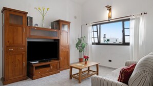 A living room with a TV, wooden cabinets, a small table, a sofa, and a window with a view. - Peaceful Villa near Famara – Ideal for Families & Groups, Up to 9 Guests (House + Studio) (Soo)