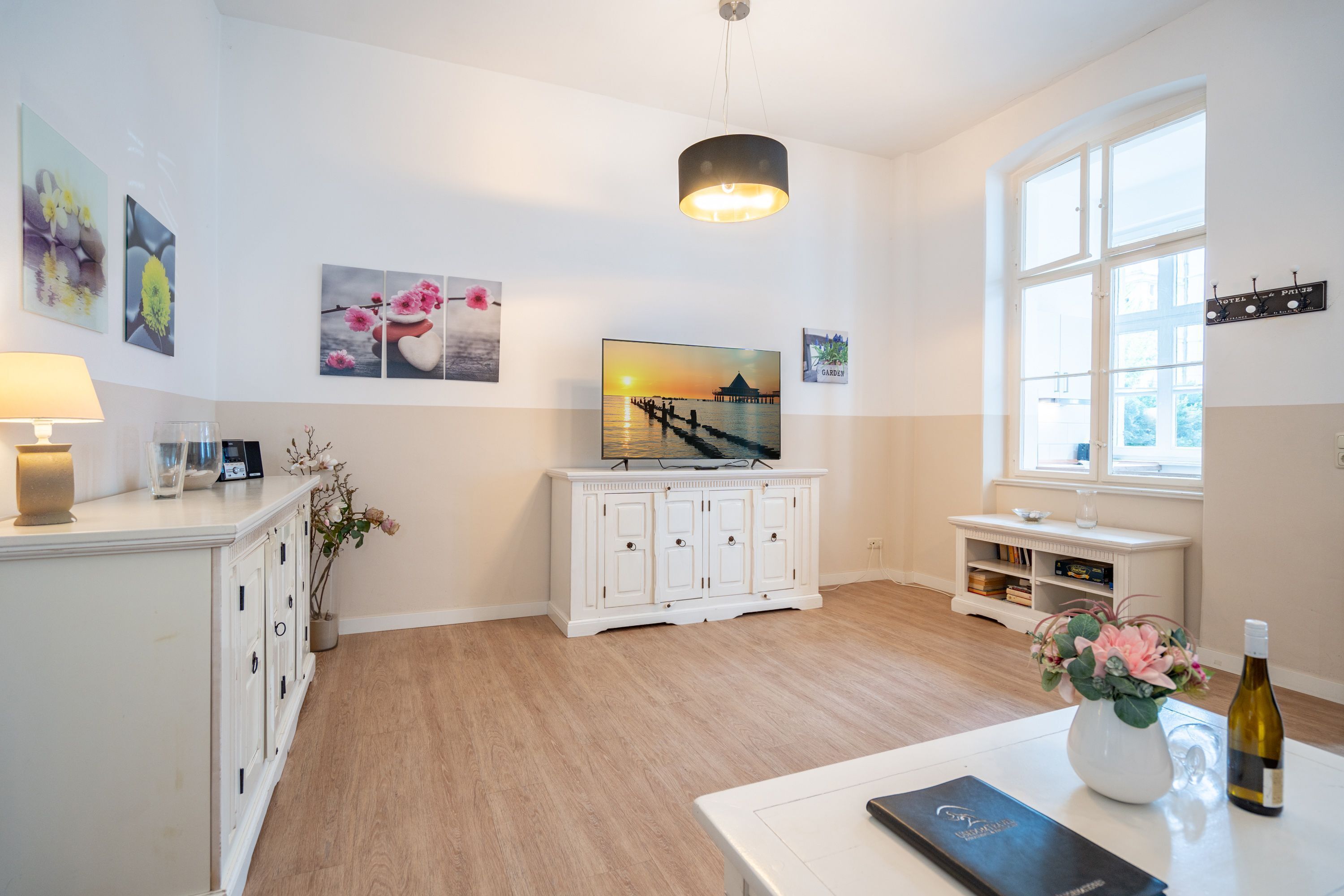 A modern living room with a white TV stand, a white cabinet, a dining table with a vase and a bottle, and a window with a view of a pier.