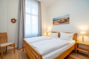 Een netjes ingerichte slaapkamer met een tweepersoonsbed, twee bedlampjes, een houten stoel, een raam met dunne gordijnen en een schilderij aan de muur.