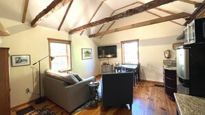 A cozy living room with a sofa, a small table, and a television. There is a kitchen area with a refrigerator and a microwave. The room has wooden beams on the ceiling and large windows.