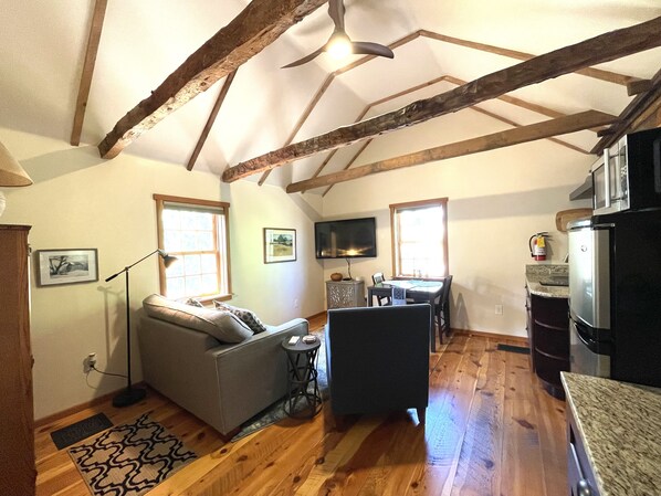A cozy living room with a sofa, a small table, and a television. There is a kitchen area with a refrigerator and a microwave. The room has wooden beams on the ceiling and large windows.