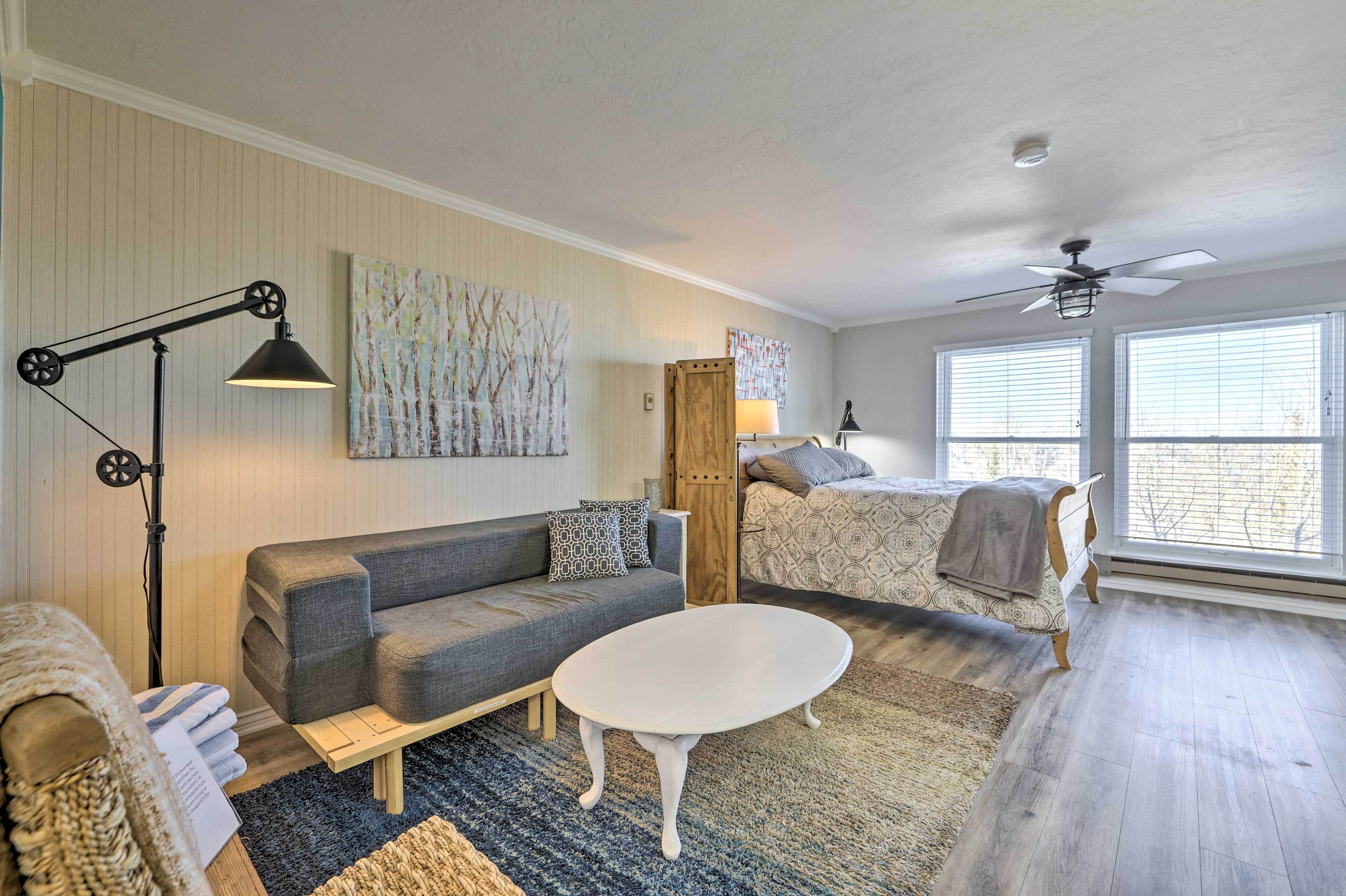 Une pièce avec un canapé gris, une table basse blanche, un lampadaire, un lit avec une literie à motifs et un ventilateur de plafond.