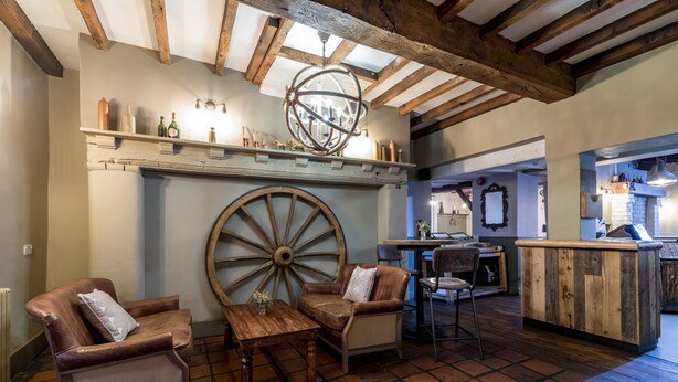 Een rustiek interieur met zichtbare houten balken, een groot houten rad, leren fauteuils, een houten tafel en een barruimte met planken.