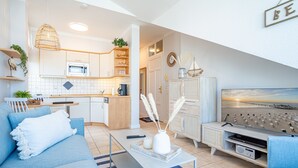 Een moderne woonkamer met een bank, salontafel en televisie. Achtergrond toont een keuken met een magnetron en kasten.