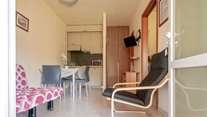 A small living area with a sofa, a chair, a dining table with chairs, a kitchen in the background, and a sliding glass door.