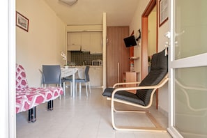 Una sala pequeña con un sofá, una silla, una mesa de comedor con sillas, una cocina al fondo y un ventanal corredero.