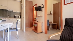 A compact kitchen with a microwave, a dining table with chairs, a small wooden shelf, and a television mounted on the wall.