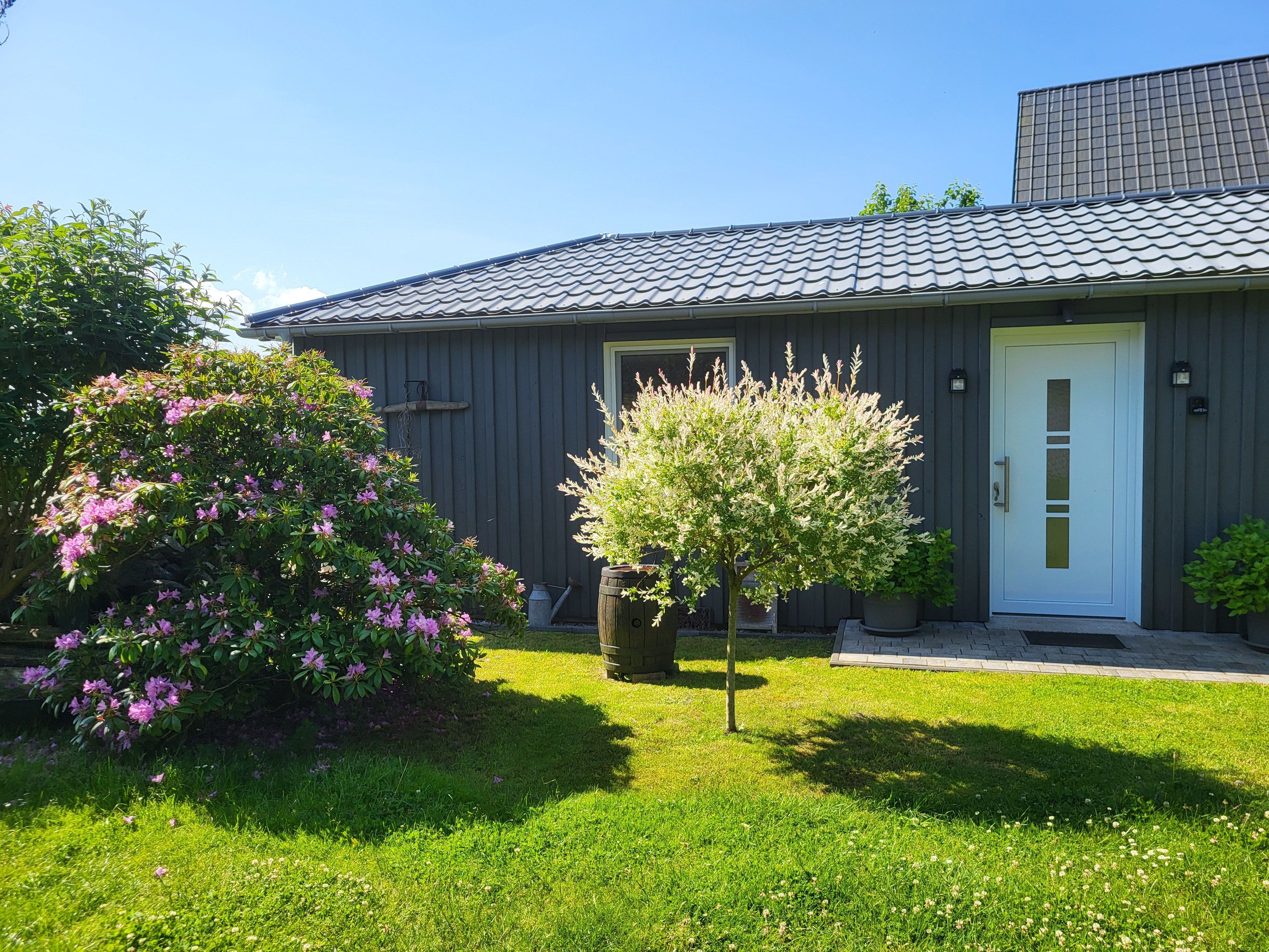 Une maison d’habitation avec un toit gris, une porte blanche et un petit arbre dans le jardin.