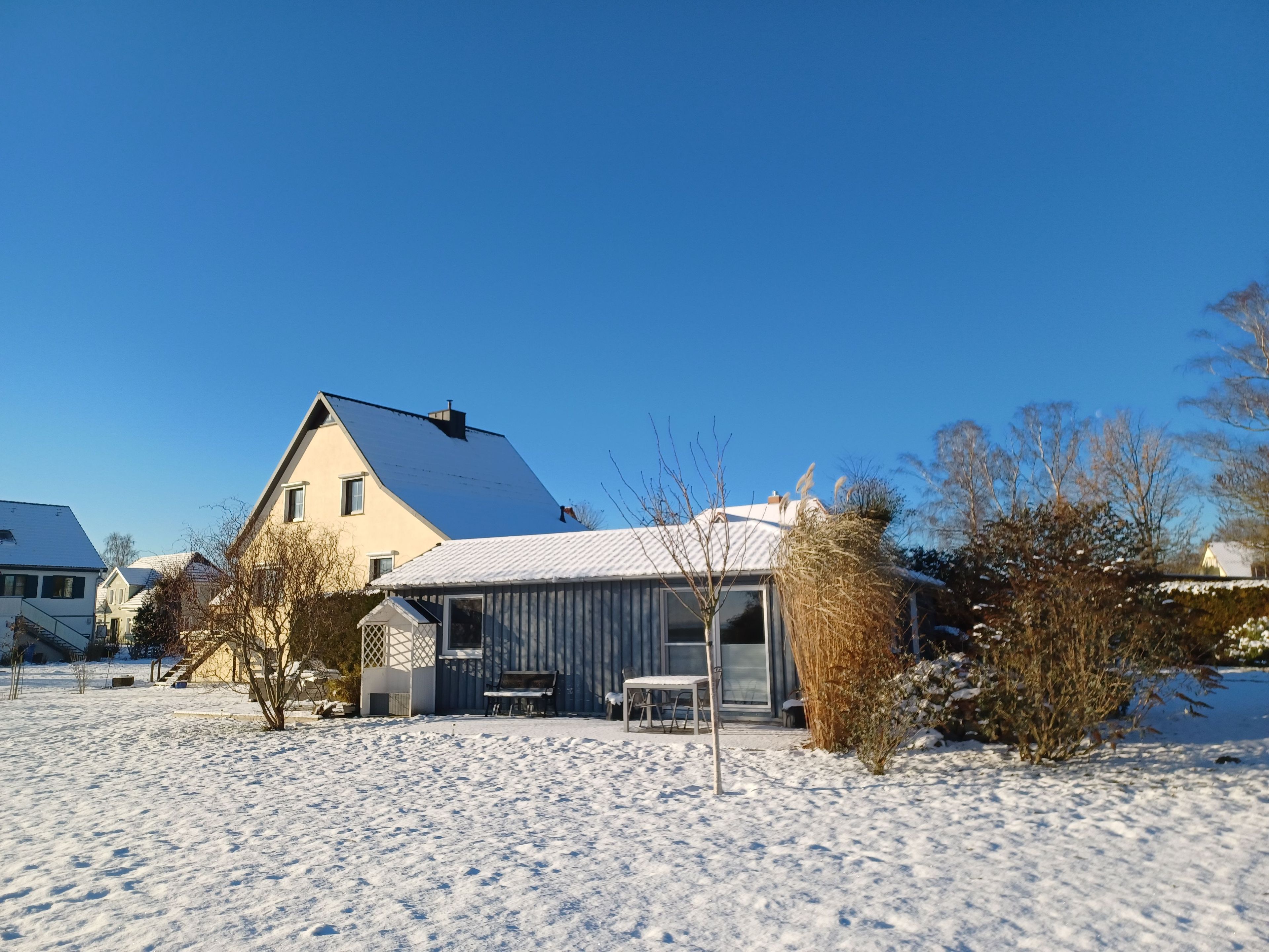 Ein Haus mit verschneitem Vorgarten, einem sichtbaren Schornstein und einem kleinen Schuppen.