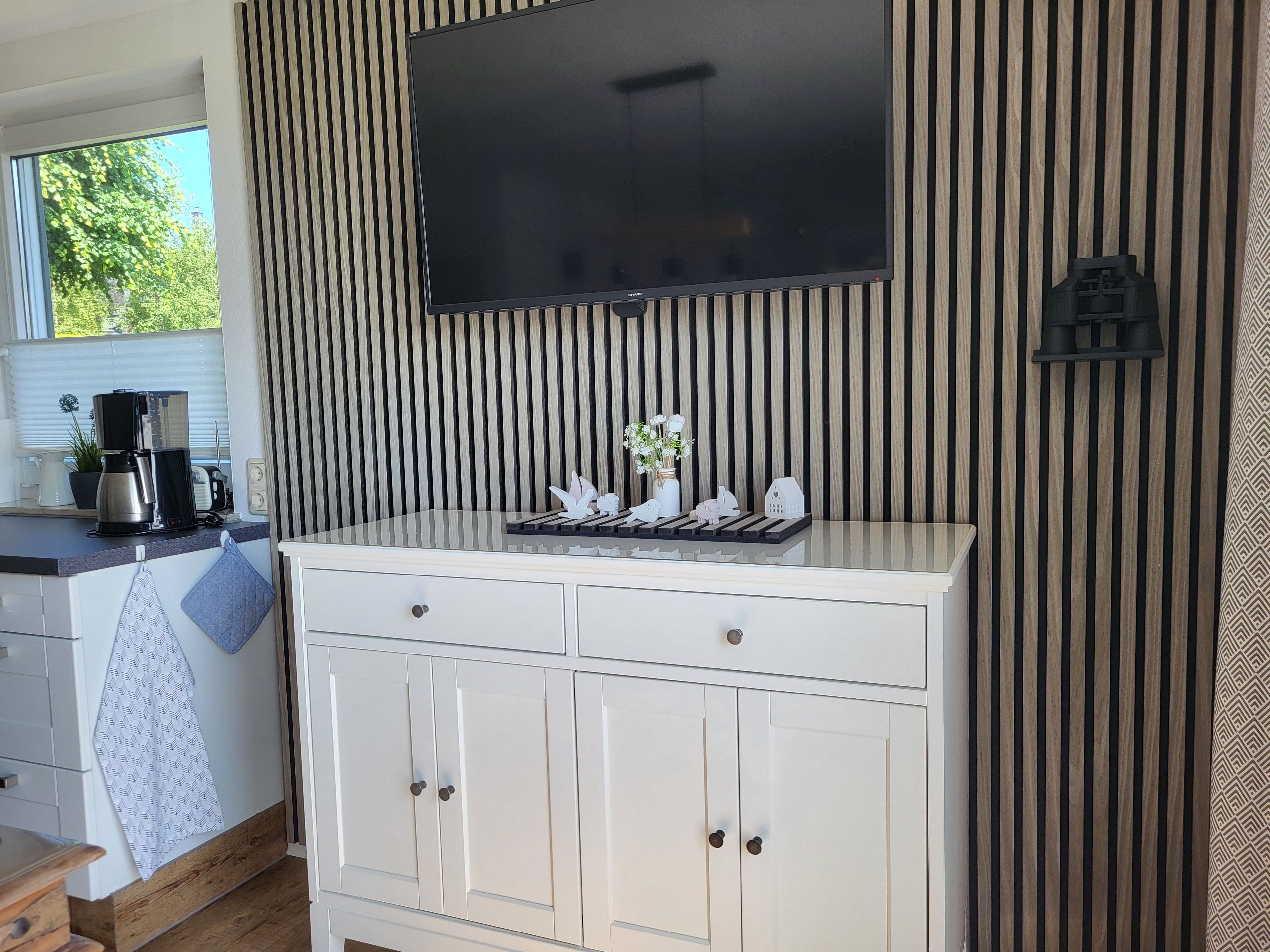 A kitchen with a TV mounted above a white cabinet, a coffee maker, and a window with blinds.