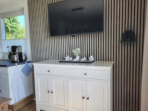 A kitchen with a TV mounted above a white cabinet, a coffee maker, and a window with blinds. - Cathrin's Boddenblick - View of Cathrin's Bodden (Wustrow)