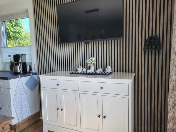 A kitchen with a TV mounted above a white cabinet, a coffee maker, and a window with blinds.