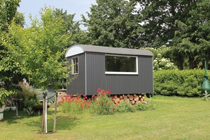A small, dark-colored cabin with a window, surrounded by a garden with red flowers and a green lawn.