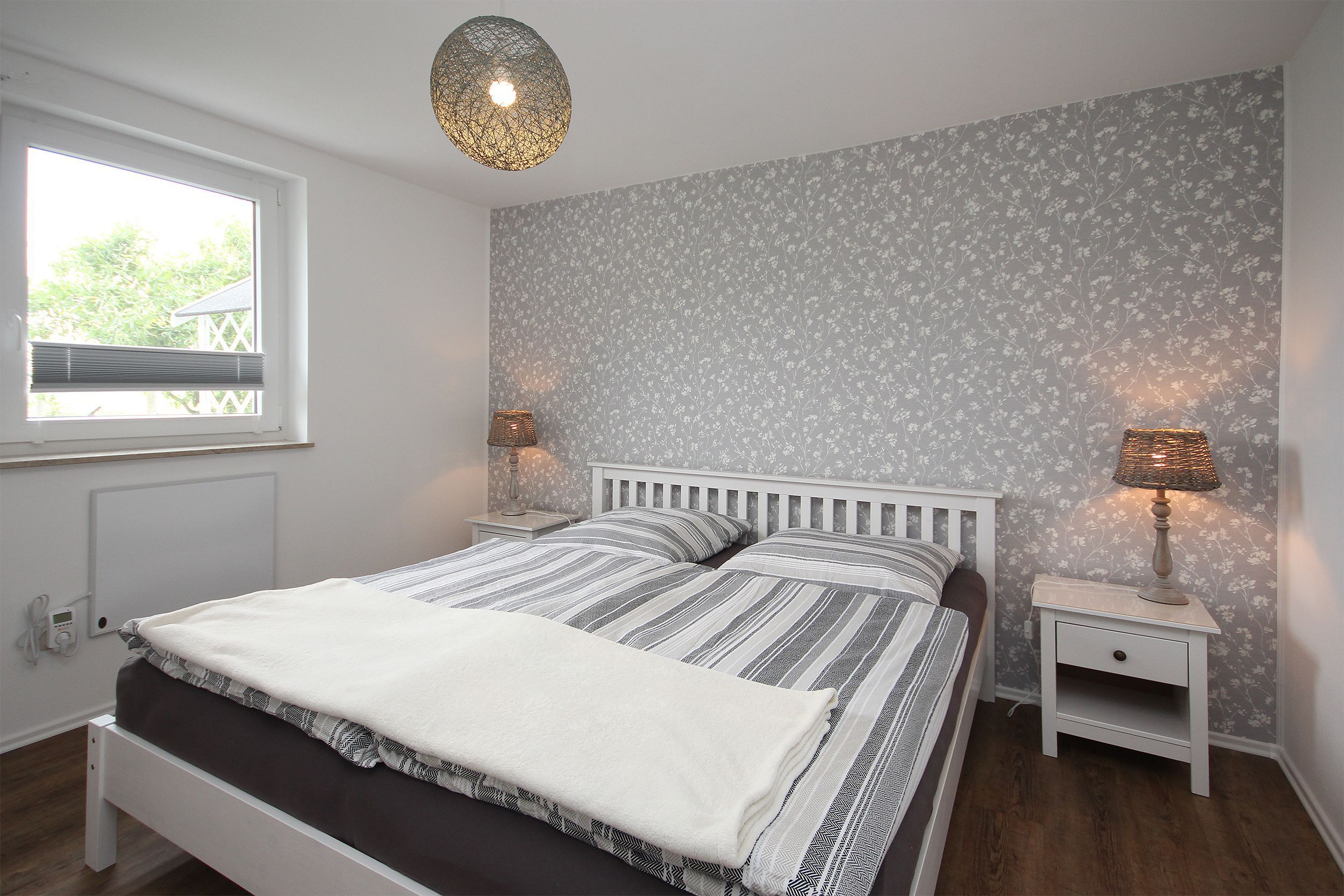 Ein Schlafzimmer mit einem Bett, Nachttischen, einem Fenster und einer Wand mit floralem Wandpapier.