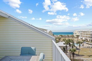 Un balcon avec vue sur la plage et l’océan, un espace piscine agrémenté de palmiers et un bâtiment à la façade jaune clair.
