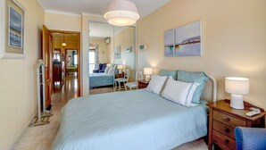 A hotel room with a large bed, bedside tables, a chair, a mirror, and a view into the hallway. - Apartment 'Beira Mar' with Sea View, Terrace & Wi-Fi (Armação de Pêra)
