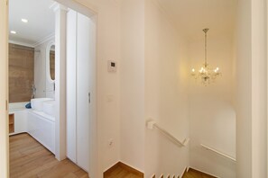 A hallway with a white wall, a wooden floor, a chandelier, and a door leading to a bathroom.