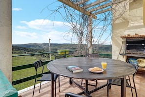 Un patio exterior con una mesa redonda, sillas, un libro, un croissant y un vaso de jugo de naranja.