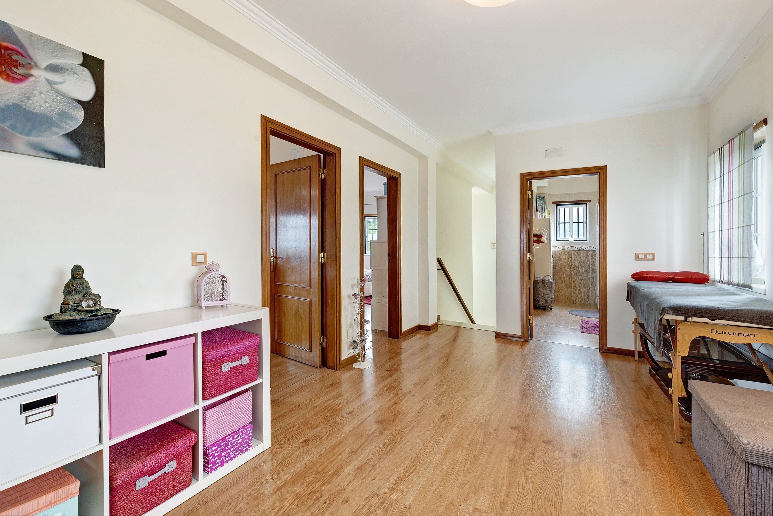 A hallway with wooden flooring, white walls, and a bed with a red pillow. There are pink storage bins and a small statue on a shelf.