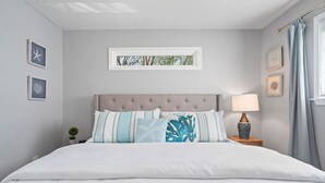 A neatly made bed with a tufted headboard, a bedside lamp, and framed artwork on the walls. - The Minted Mermaid - Pet Friendly - Winter Texan Rates! (Galveston)