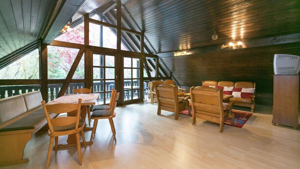 A spacious room with a high ceiling, wooden beams, a large dining table with chairs, a sofa set, and a refrigerator.