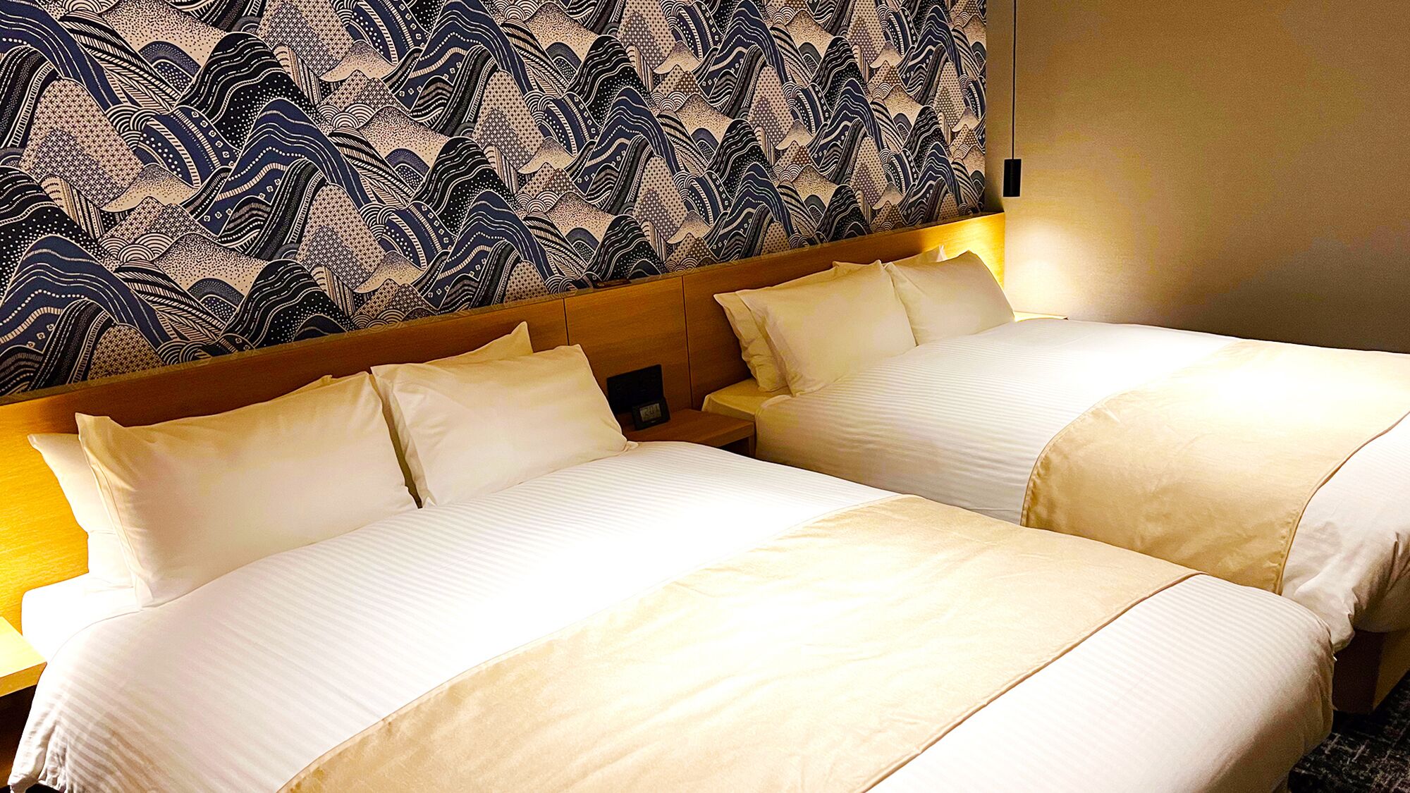 A hotel room with two beds, a headboard with a patterned wallpaper, and a bedside table with a lamp.