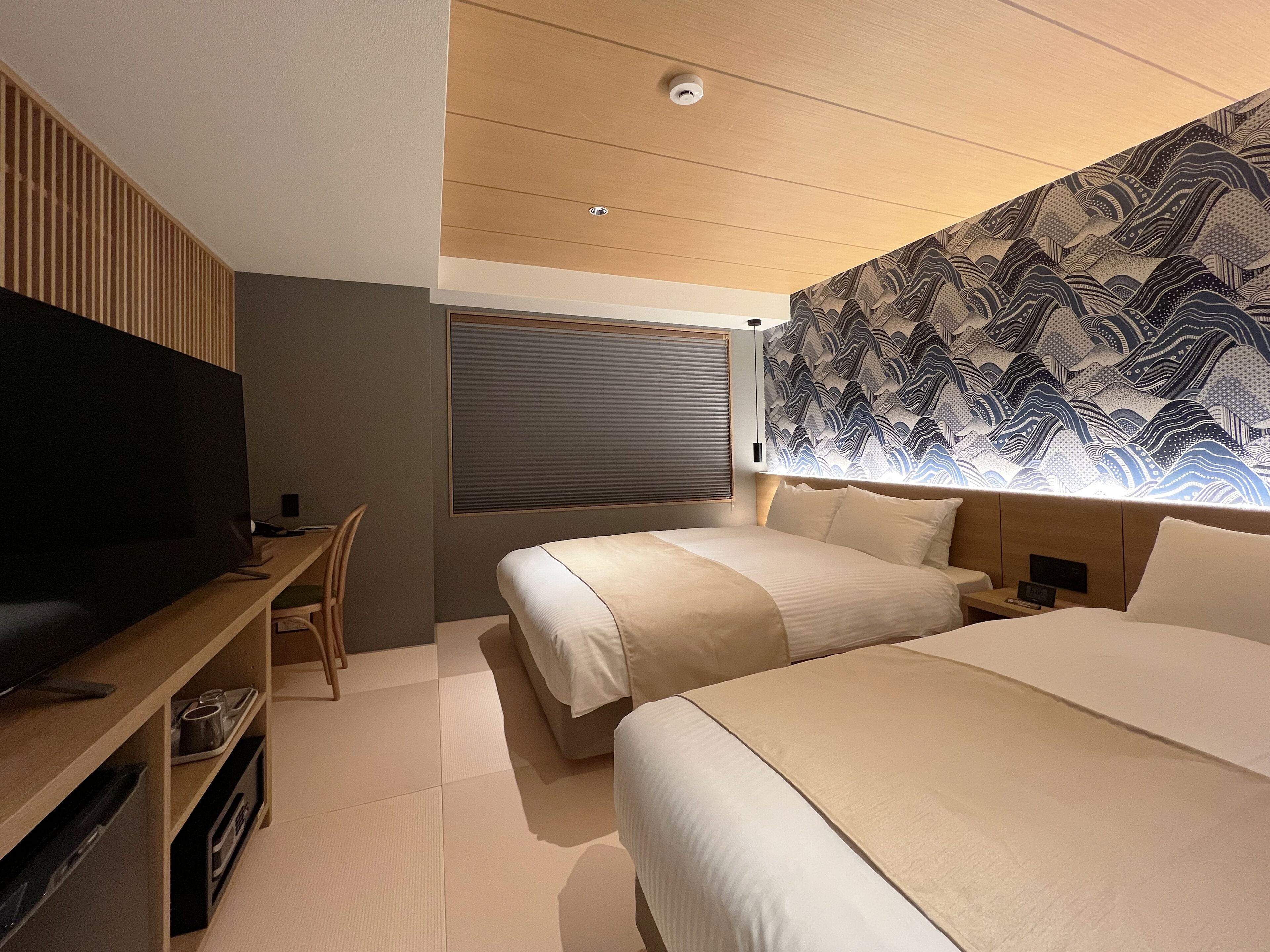 A modern hotel room with a large flat-screen TV, two beds, a desk with a chair, and a patterned wall.