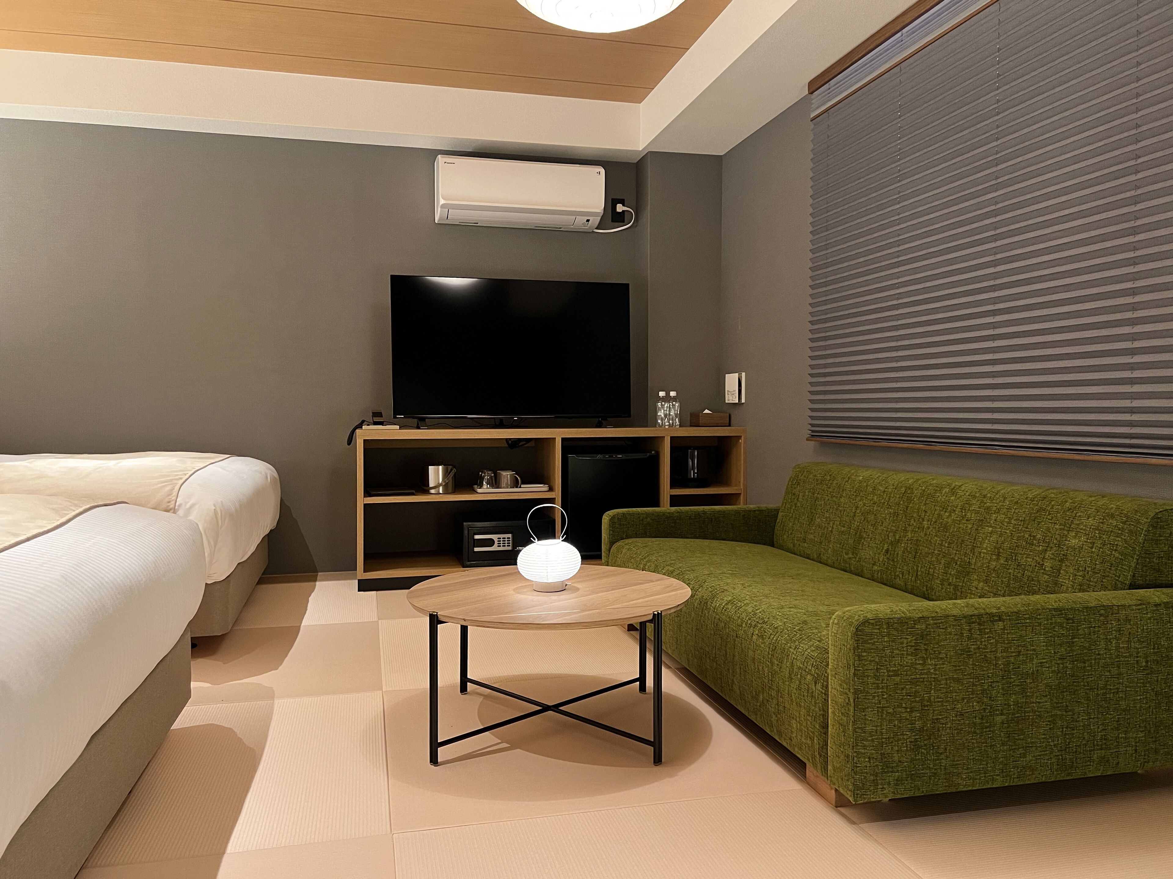 Una habitación de hotel moderna con una cama, un sofá verde, una mesita redonda pequeña, un televisor de pantalla plana y un equipo de aire acondicionado integrado.