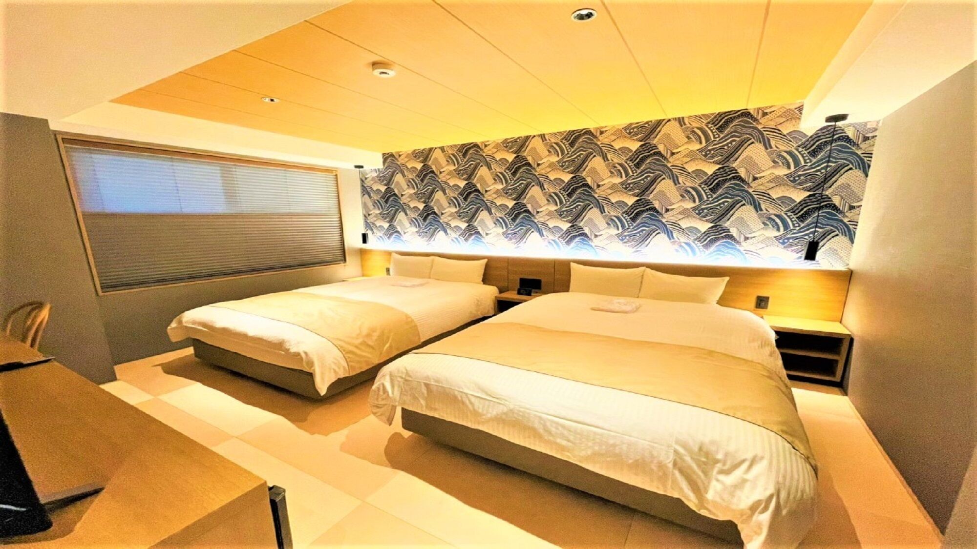 A bedroom with two beds, a window with blinds, a patterned wall, and a ceiling light fixture.