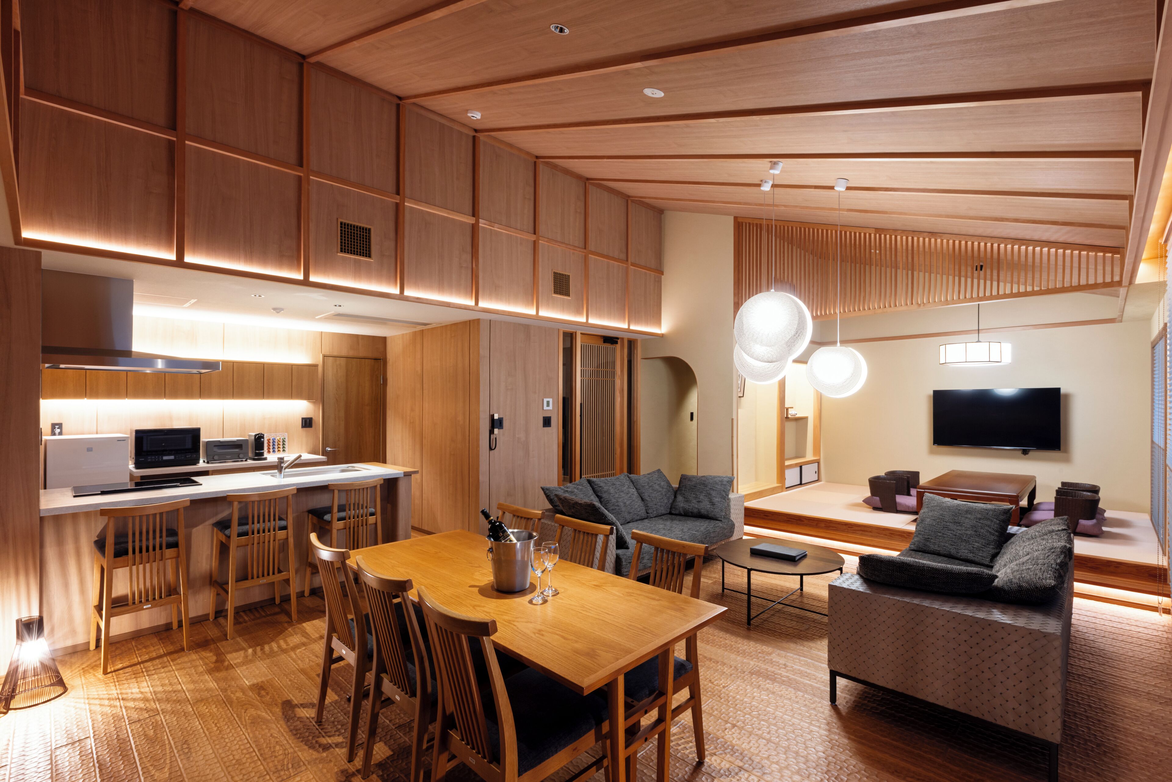 A modern kitchen with wooden cabinetry, a dining area with a wooden table and chairs, a sofa, and a flat-screen TV mounted on the wall.