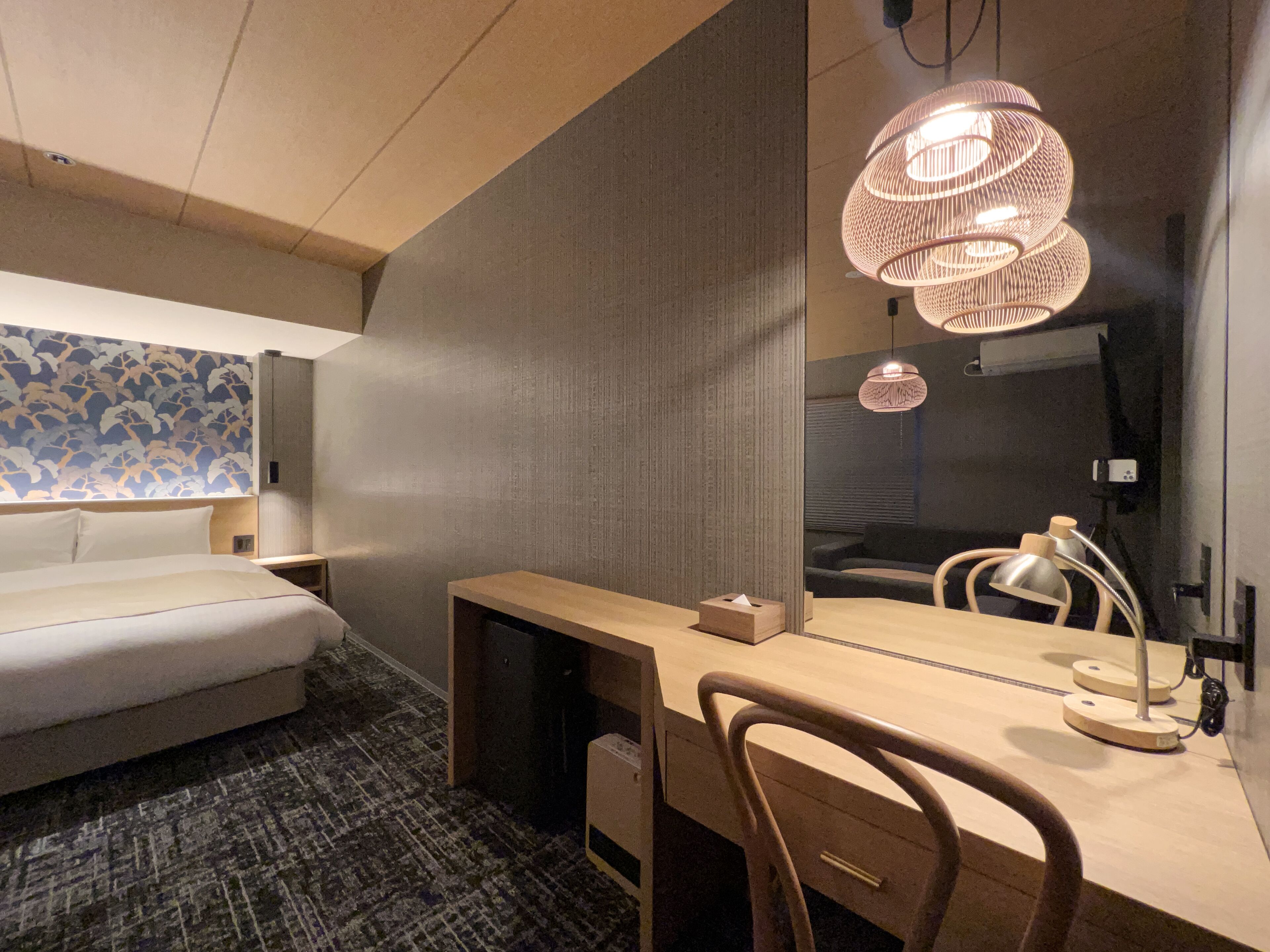 Une chambre d’hôtel moderne équipée d’un lit, d’un bureau, d’une chaise, d’un miroir et d’un luminaire original en forme de sculpture tissée.