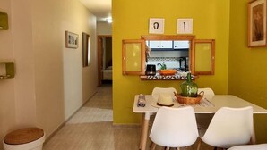 A dining area with a table set for two, a kitchen in the background, and a yellow wall.