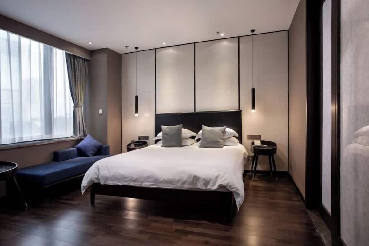 A modern hotel room with a large bed, bedside tables, a seating area, and a large window with curtains.