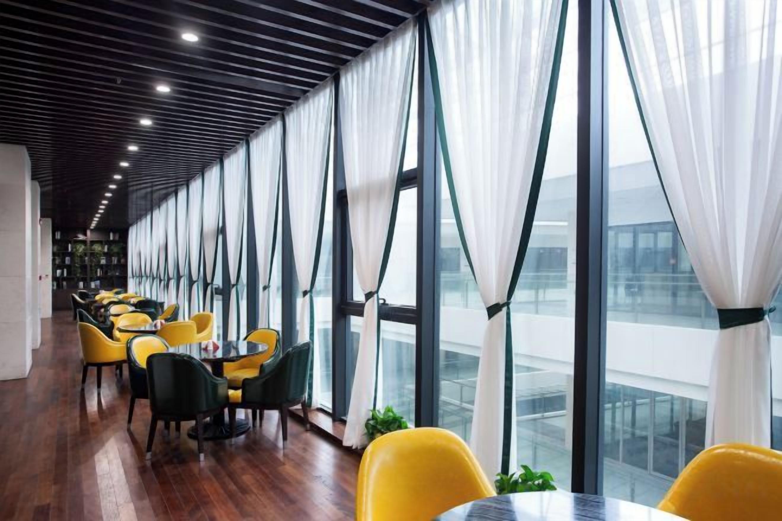 A modern interior with a long row of tables and chairs, large windows with white curtains, and a wooden floor.
