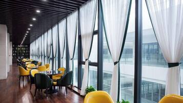 A modern interior with a long row of tables and chairs, large windows with white curtains, and a wooden floor.