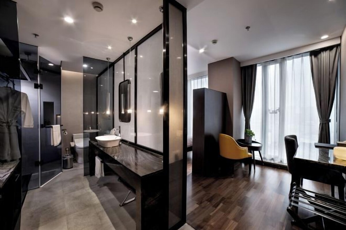 Una habitación de hotel moderna con un separador de vidrio para el baño, un espejo grande, un tocador con marco negro, suelo de madera y un área de comedor con una silla amarilla.
