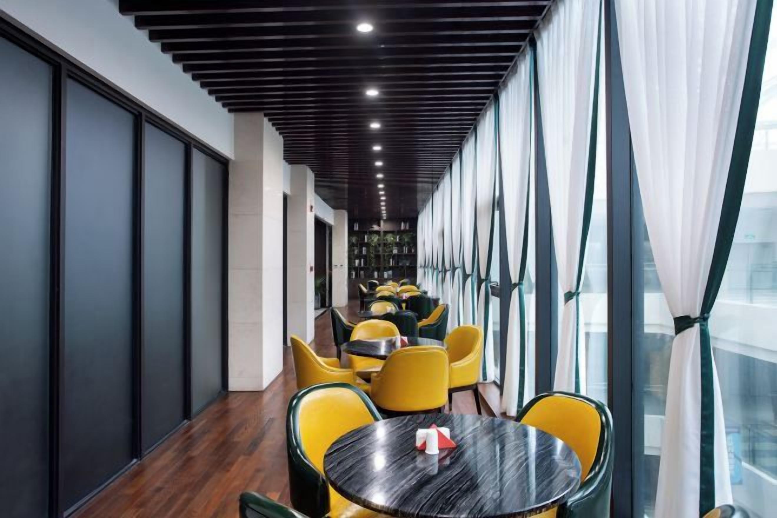 A corridor with yellow chairs and round tables, leading to a room with large windows.