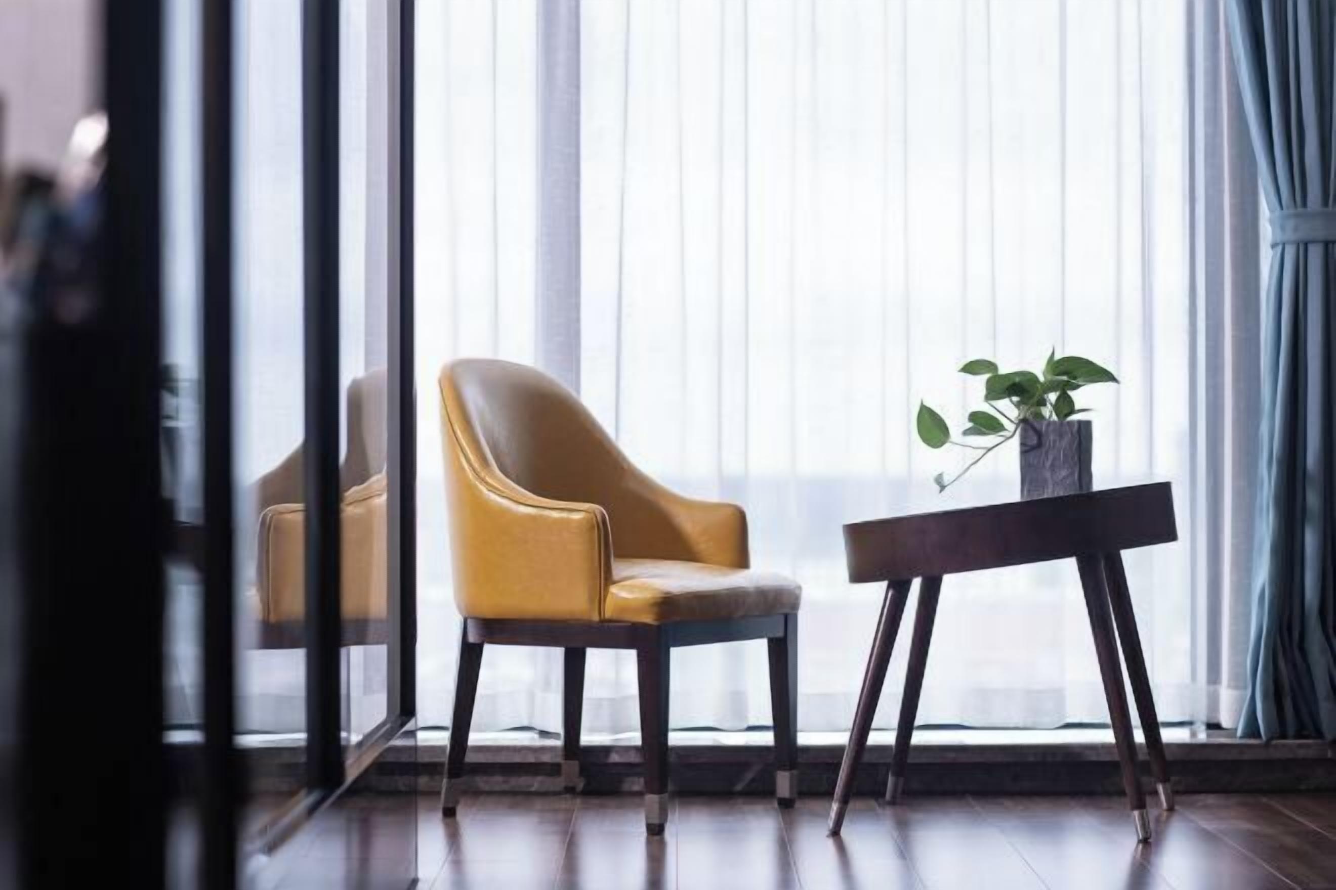 Una habitación con un ventanal grande, una silla amarilla, una mesita con una planta y suelo de madera.