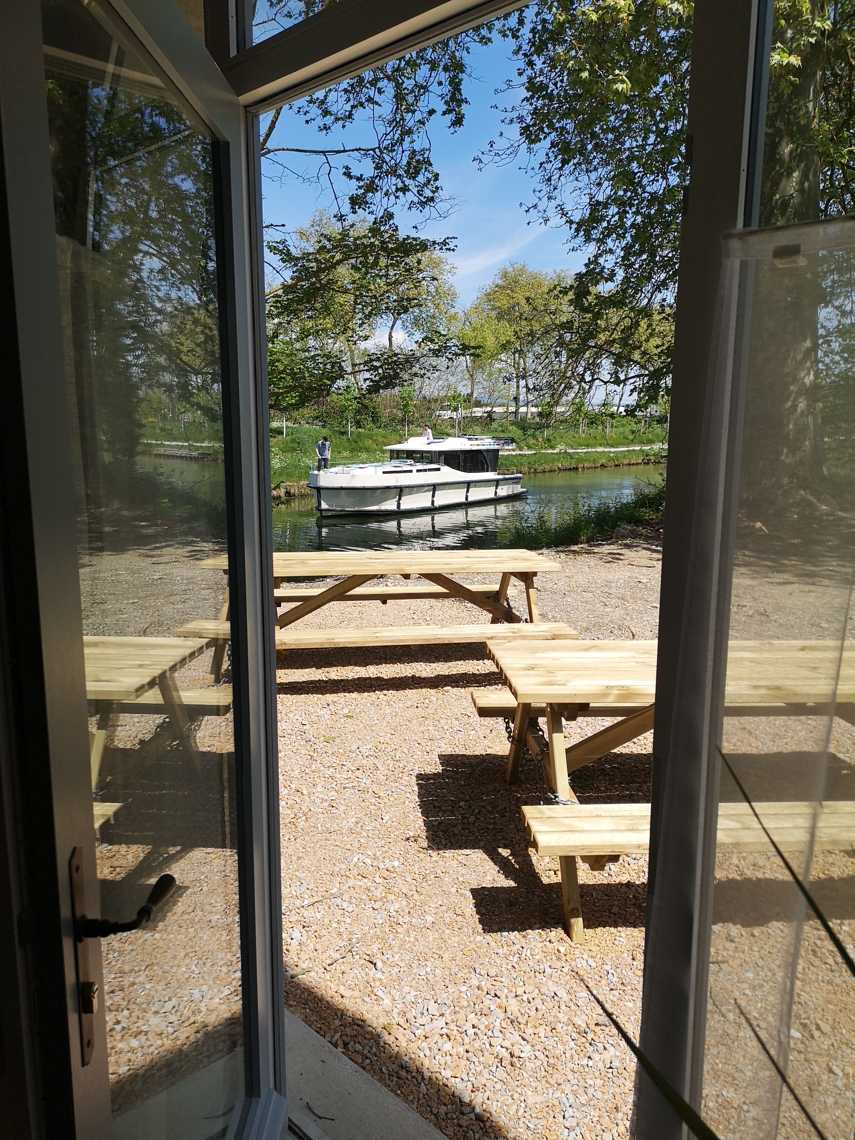A view through a glass door of a picnic area with wooden tables and benches, a boat on a lake, and trees in the background.