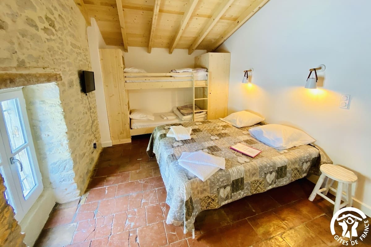 Une chambre rustique avec un lit superposé, un lit simple, un petit tabouret et un mur en pierre.