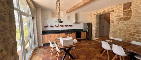 Une cuisine spacieuse aux murs en pierre, une grande salle à manger et une vue dégagée sur l’extérieur grâce à une porte vitrée.
