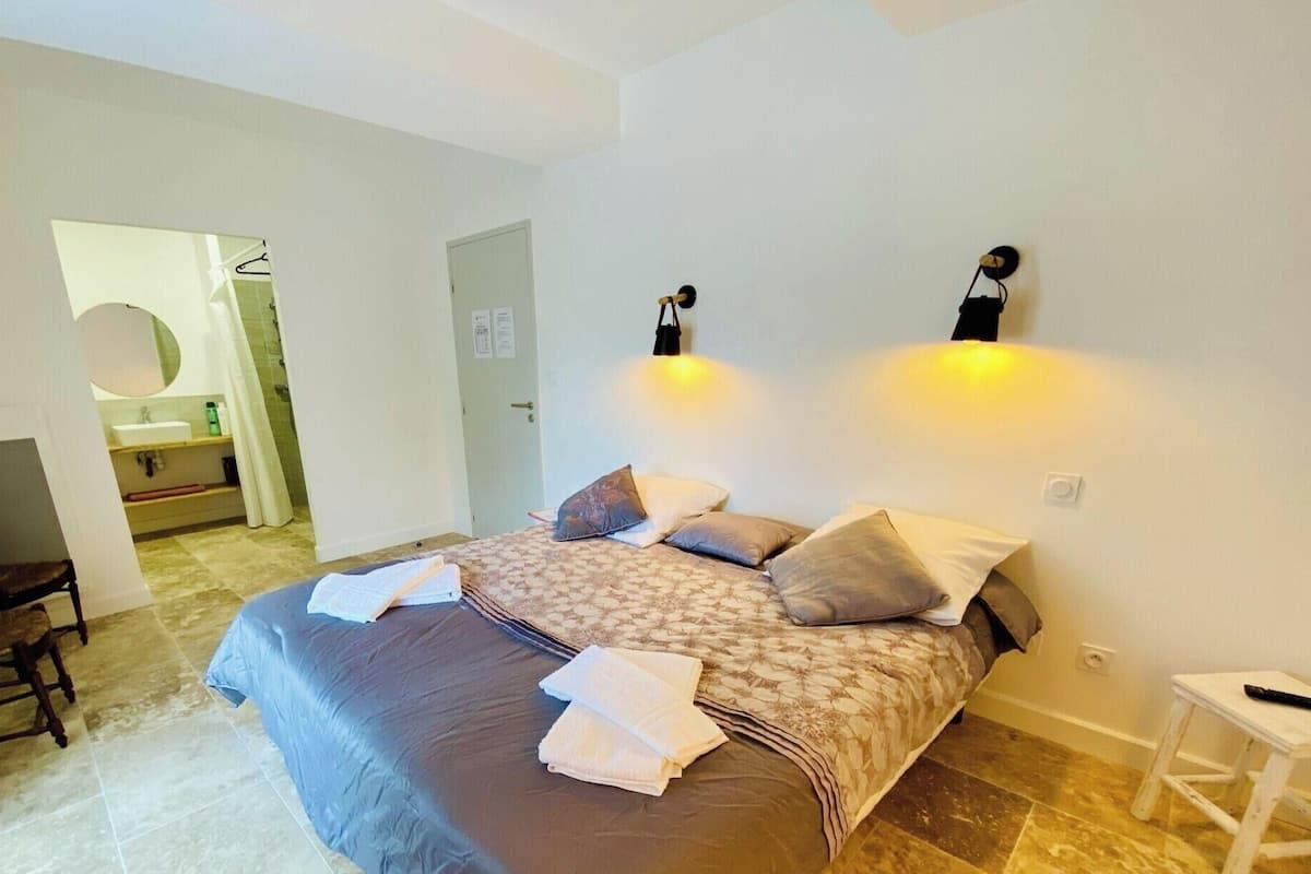 Une chambre à coucher avec un lit, des oreillers, une chaise, une petite table et un luminaire fixé au mur.