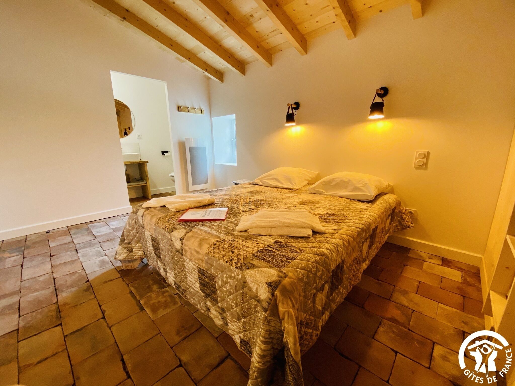 A bedroom with a large bed, wooden beams on the ceiling, and two wall-mounted lights.