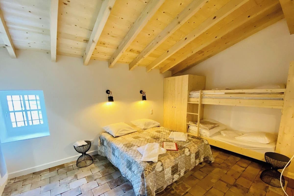 Une chambre avec un lit superposé, une petite table de chevet, une lampe fixée au mur et une fenêtre avec des stores.