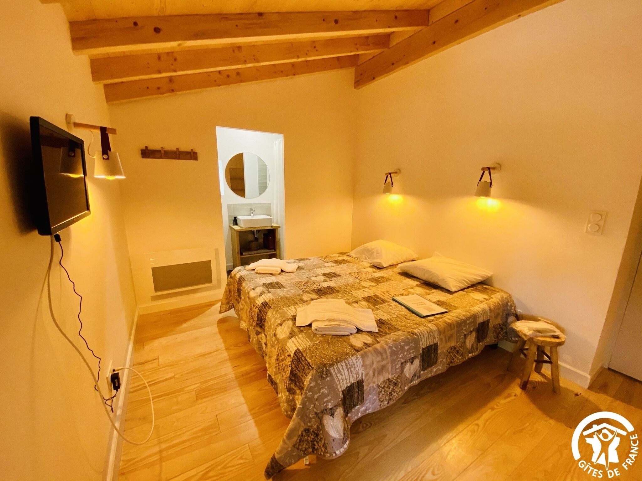A bedroom with a wooden ceiling, a bed with a patterned bedspread, a bedside table, a mirror, and a TV mounted on the wall.