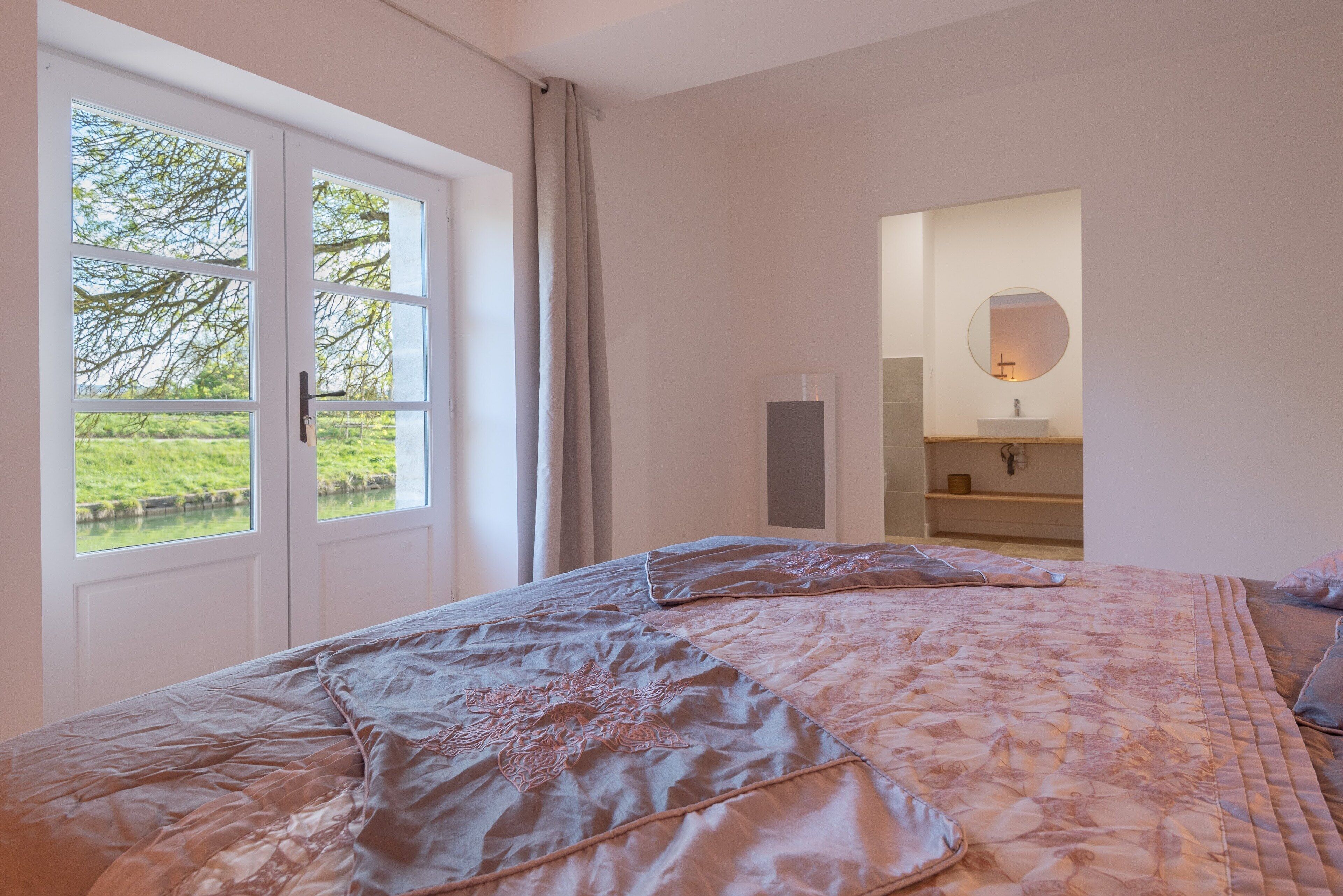 Un dormitorio con una cama grande, una ventana con cortinas y un baño con espejo y lavamanos.