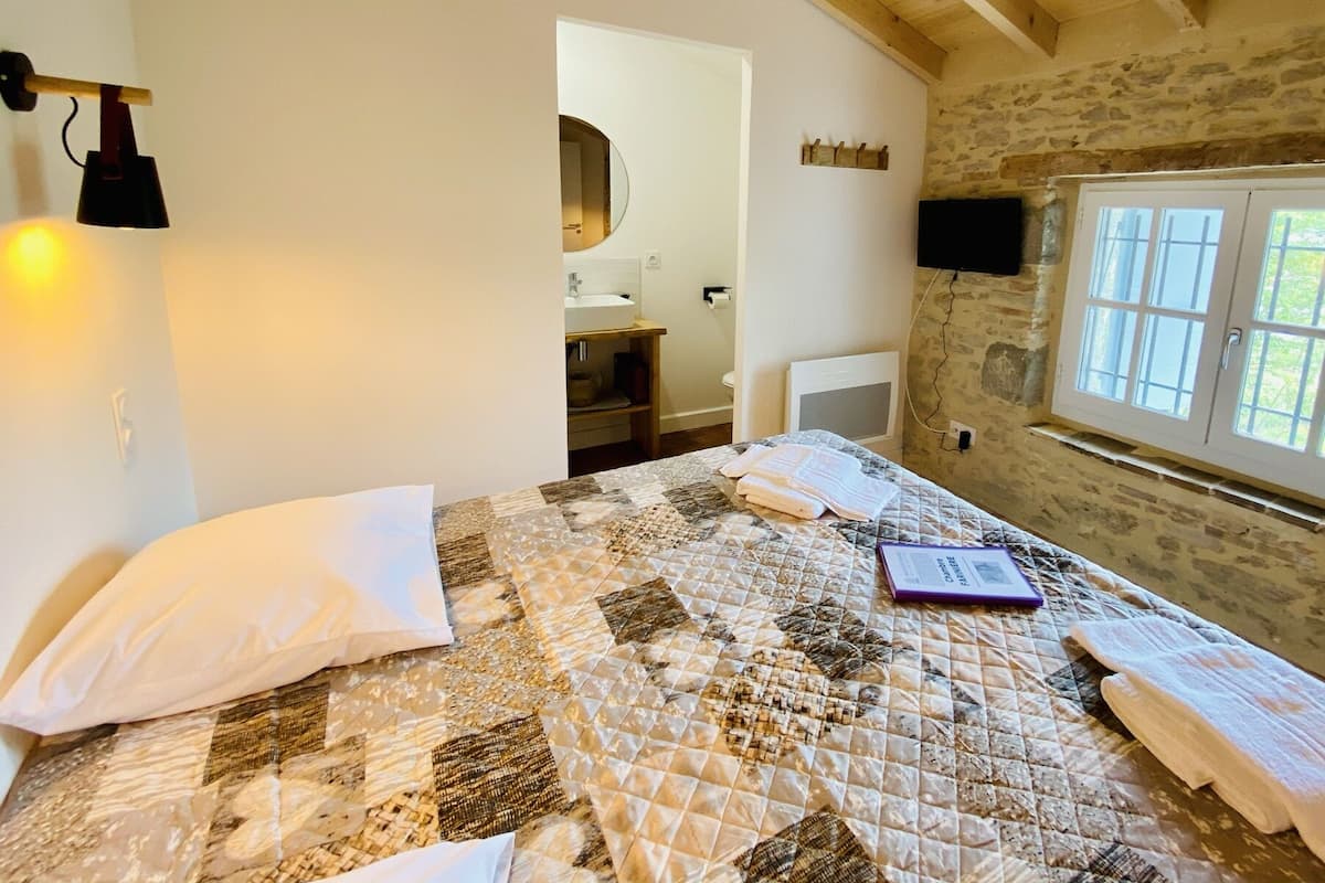 Une chambre avec une couvre-lit matelassé, une table de chevet sur laquelle repose un livre, une fenêtre avec des murs en pierre et un plafond en bois.