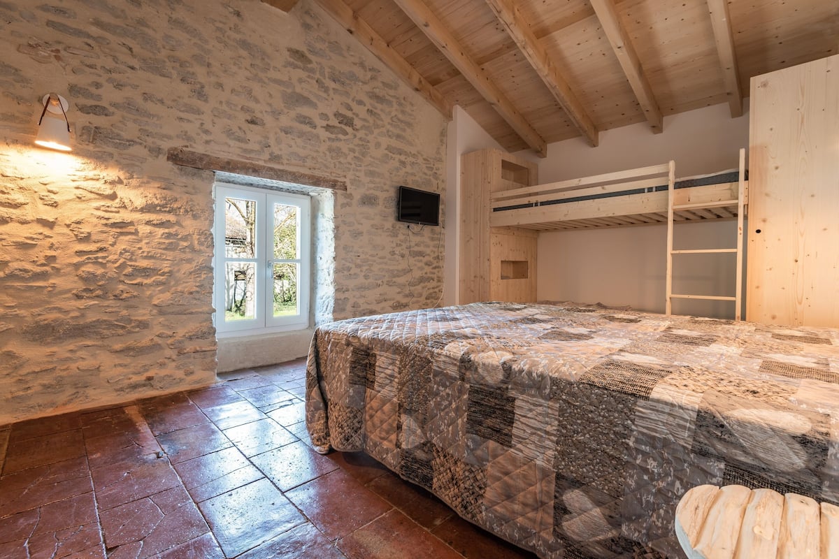 Une chambre rustique avec des murs en pierre, un plafond en bois, un lit recouvert d’une courtepointe, un lit superposé, une télévision et une porte donnant sur l’extérieur.