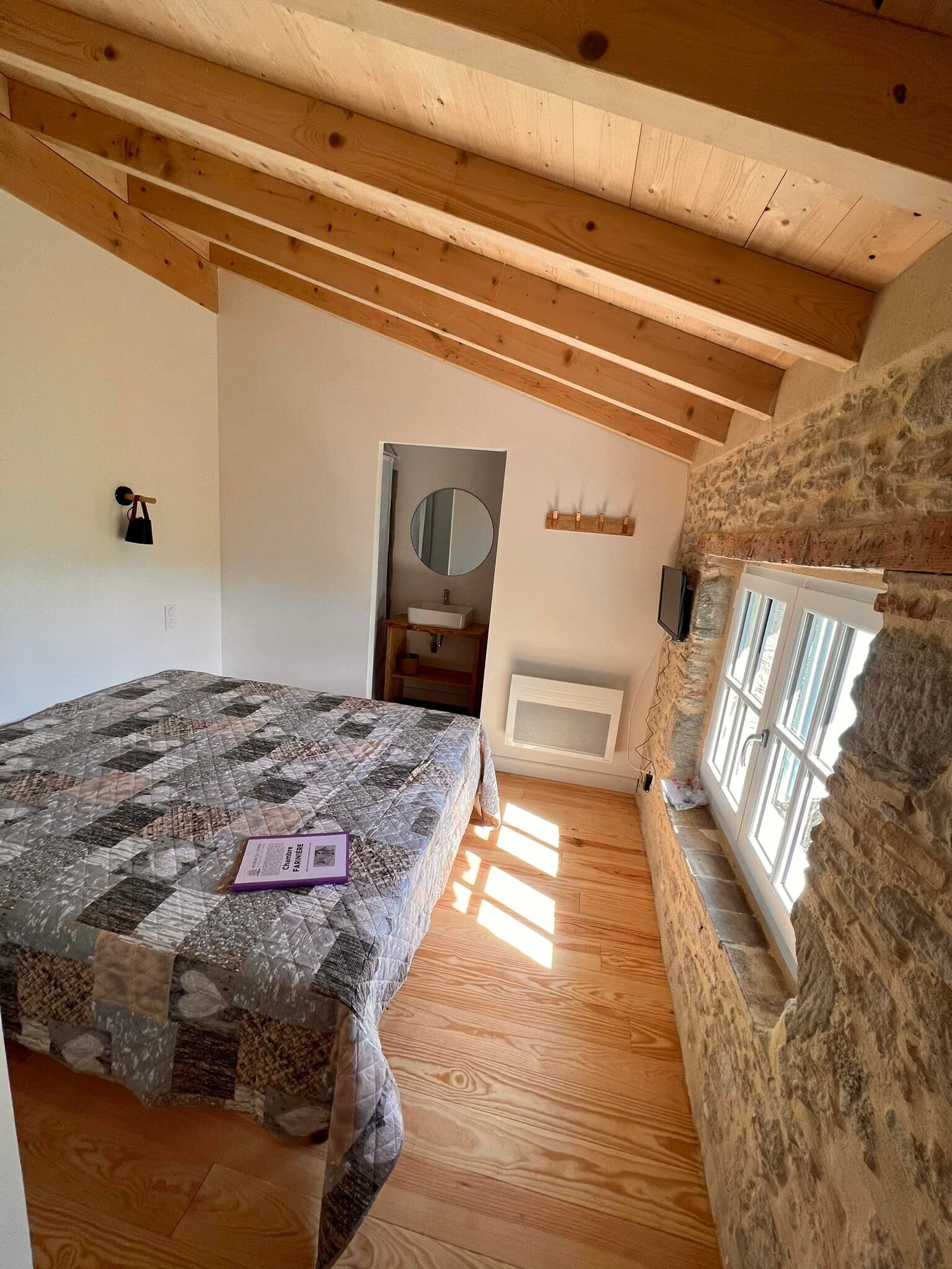 Une chambre avec un plafond en bois, un mur en pierre, un lit recouvert d’une courtepointe, une table de chevet, un miroir et une fenêtre.