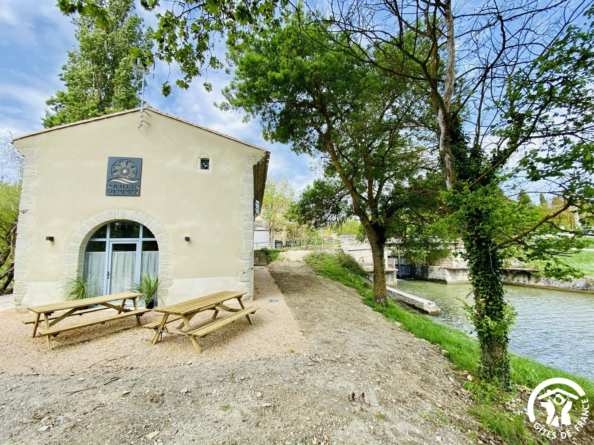 Un petit bâtiment avec une enseigne indiquant « MCULIN DU VIVIEN », situé près d’une rivière, avec une table de pique-nique et des arbres tout autour.